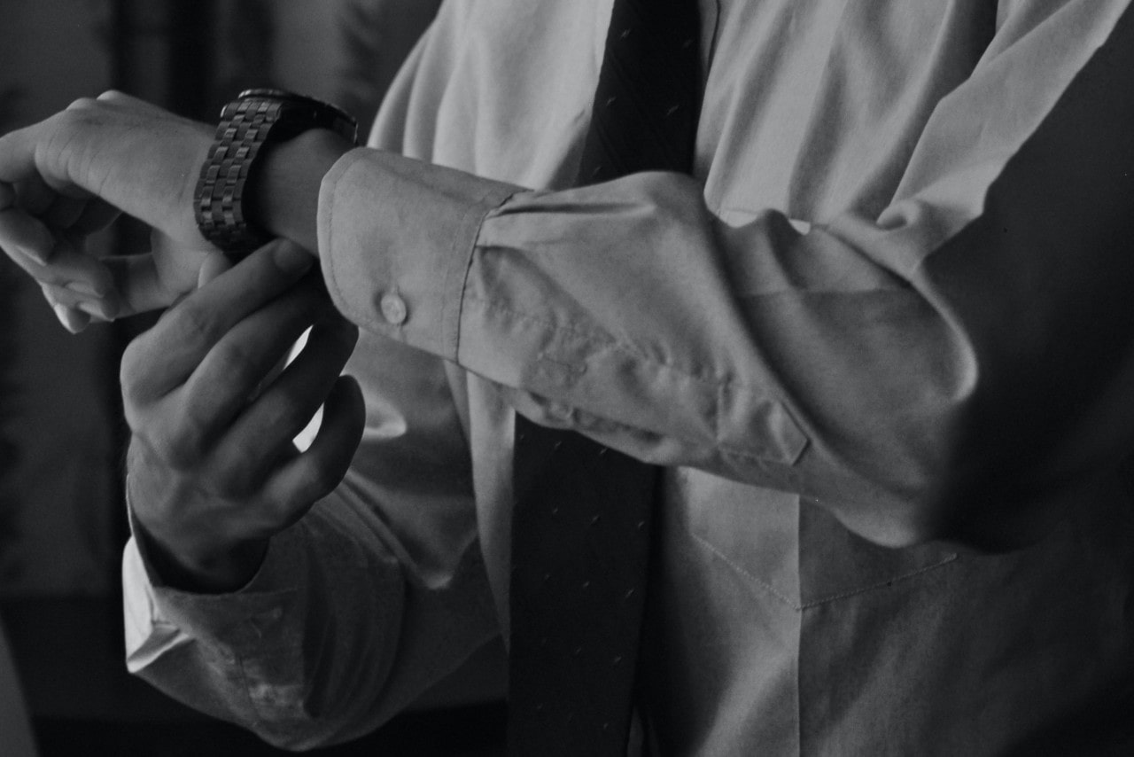 A man in a dress shirt and tie putting on his luxury watch that has a stainless steel watch bracelet and casing.