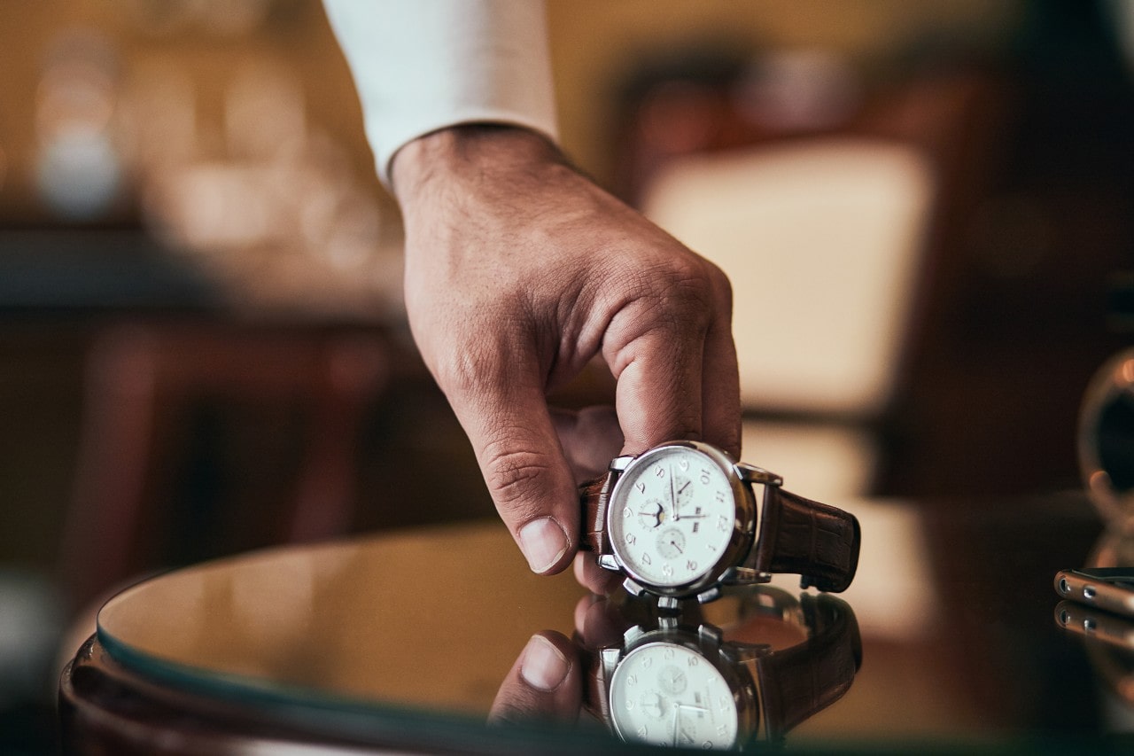 A well-dressed man picking up a luxury watch from a side table.