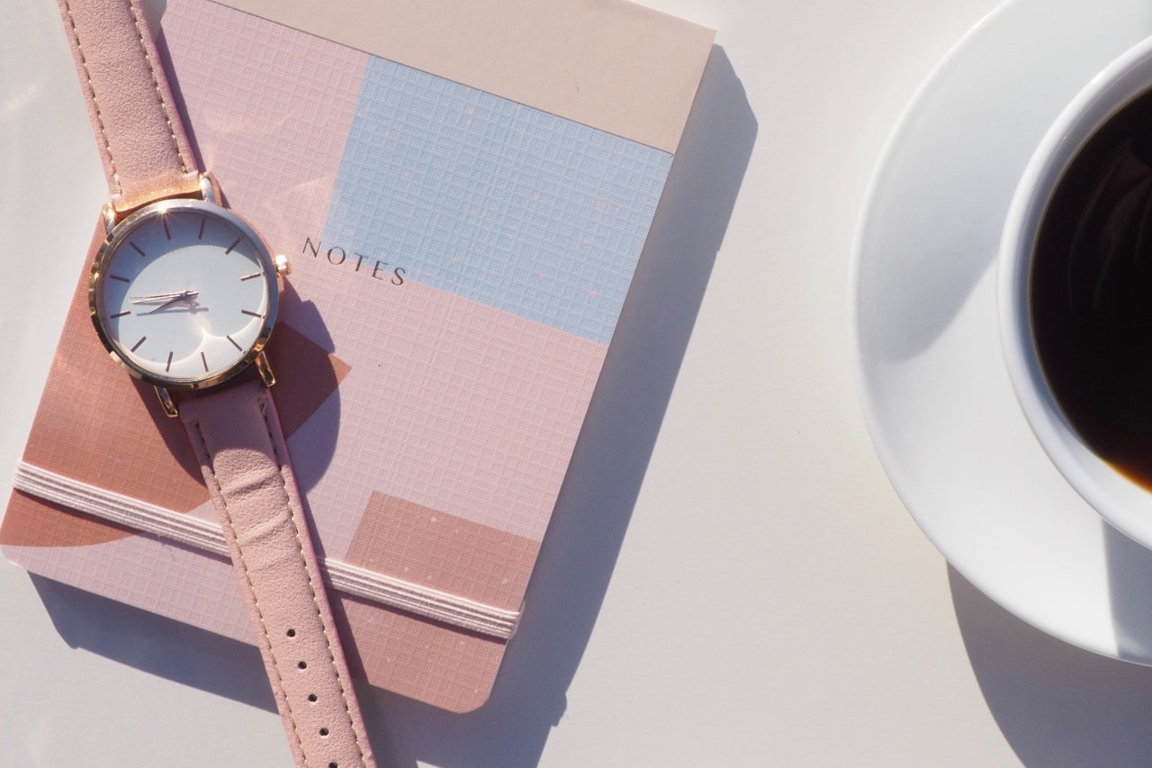 A minimalist women’s watch displayed atop a notebook next to a cup of coffee.