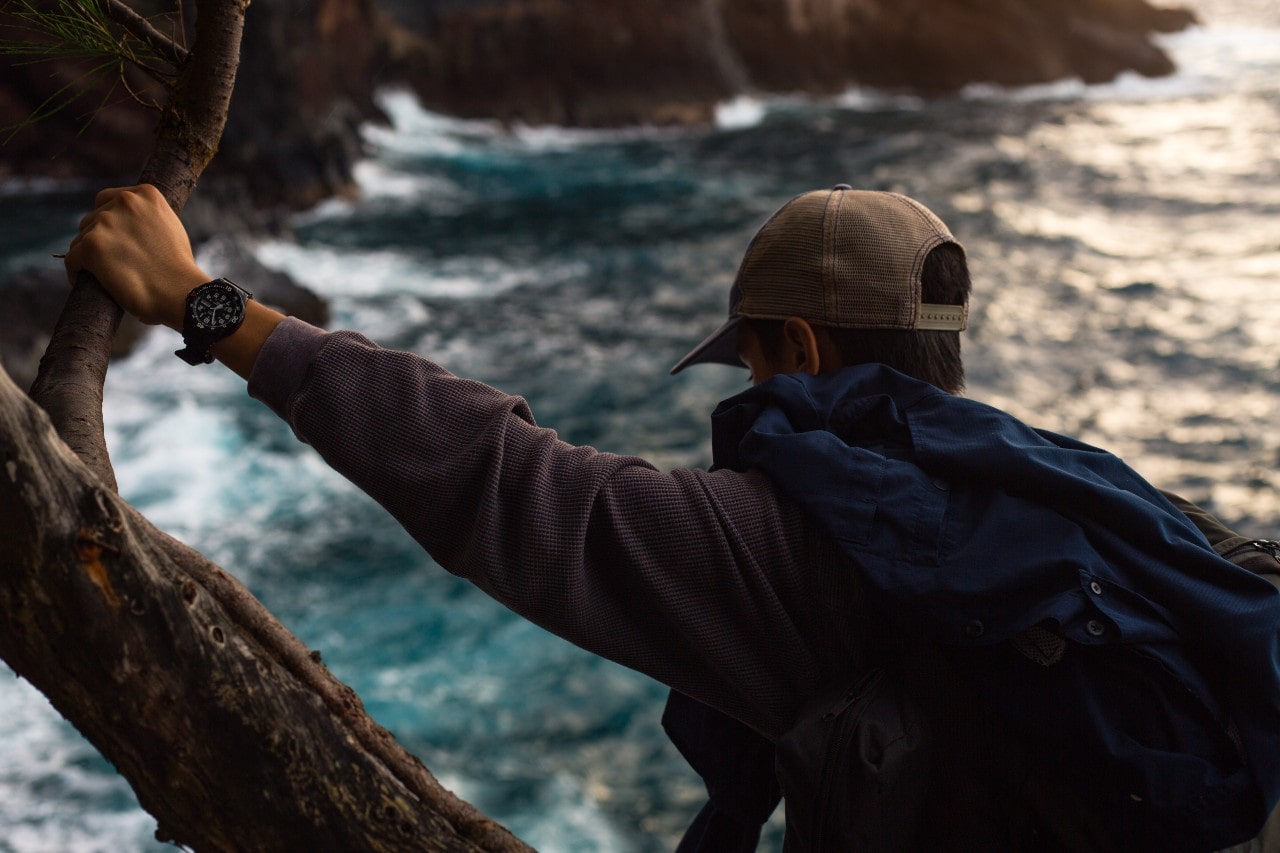 A man on a hike, stopping for a moment on a cliff,  and his watch is in view as he leans against a tree.