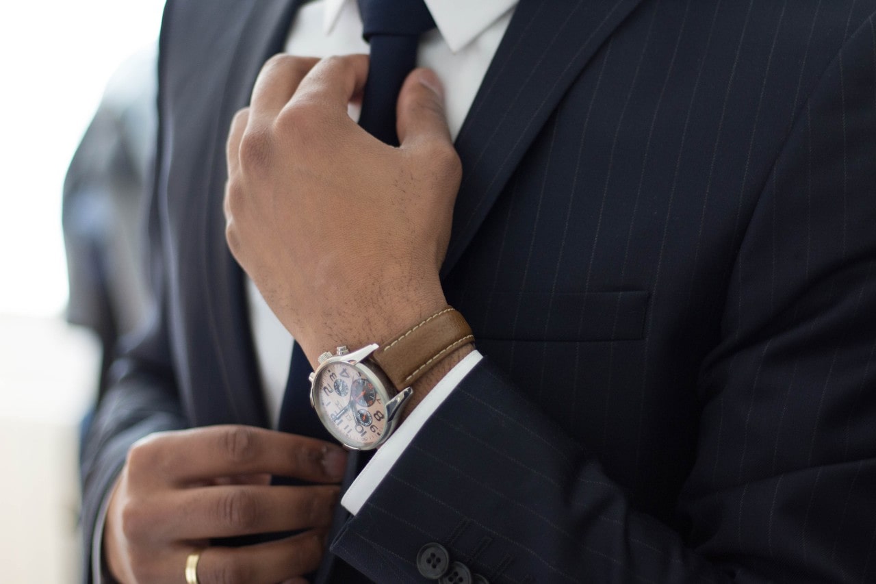 A man in a dark pinstripe suit adjusting his tie, showcasing a brown leather-strap wristwatch with a silver dial.