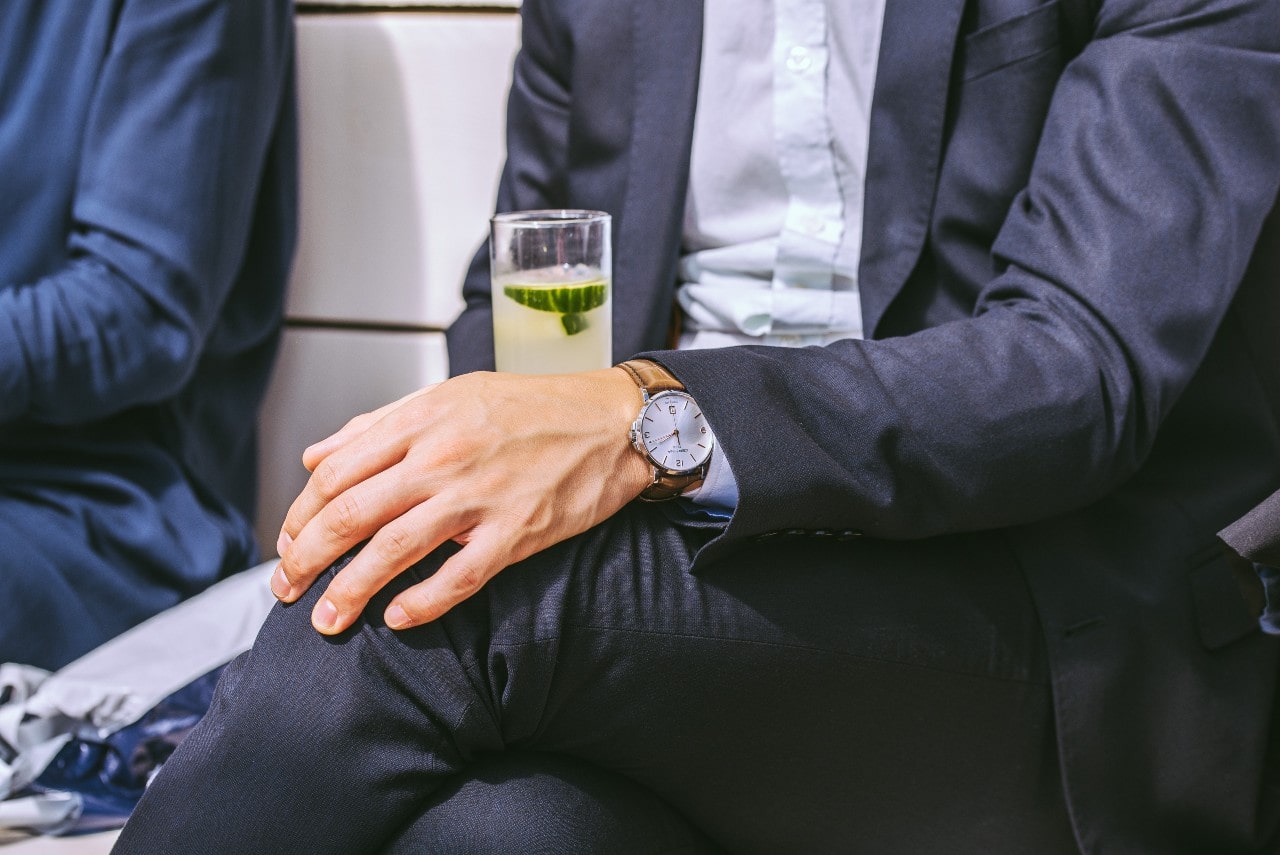 A man in a navy-blue suit sitting with his hands crossed, wearing a stylish wristwatch with leather strap and holding a drink with a cucumber garnish.