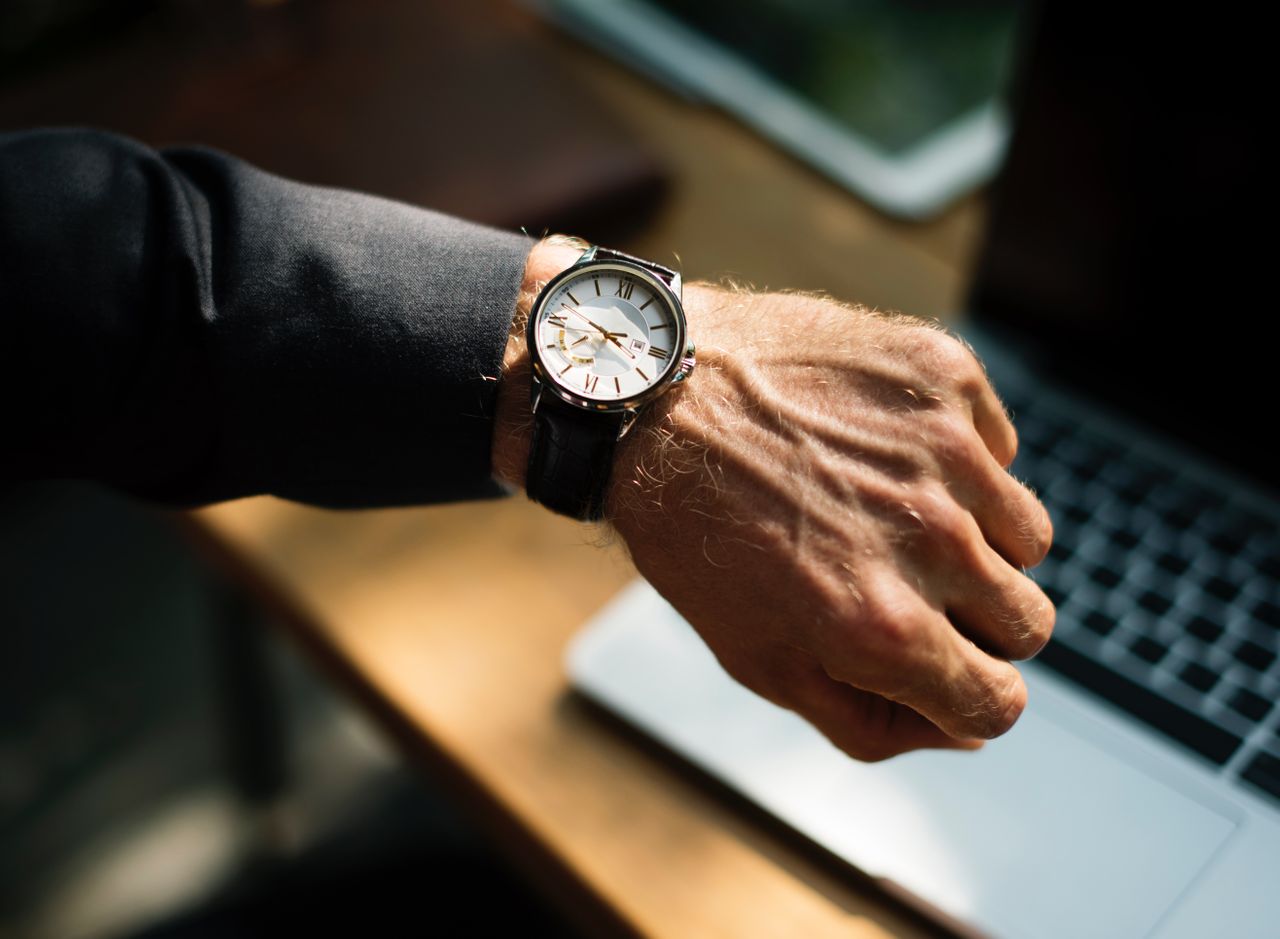 A close-up of a man's wrist with a silver watch with a white dial, Roman numeral markers, and a black leather strap.