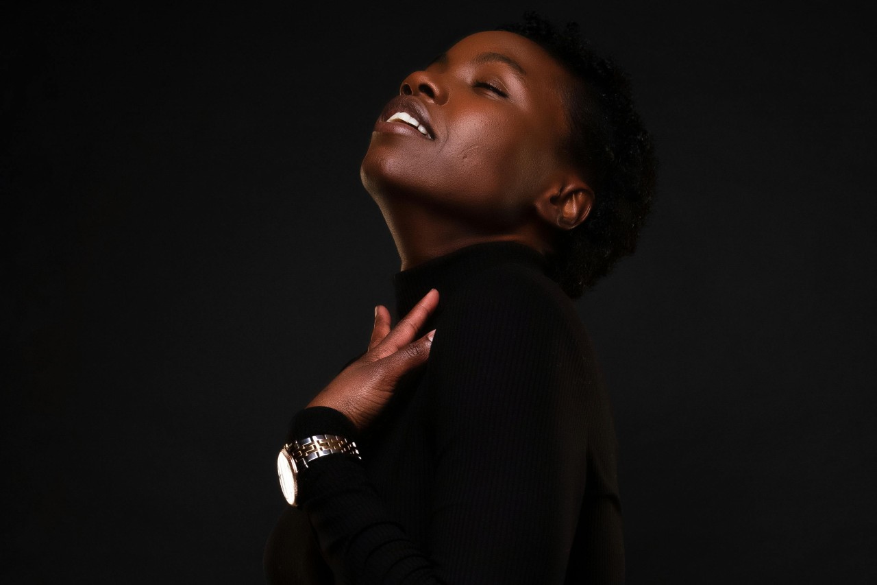 a woman in a black blouse against a black background touching her chest and wearing a bold watch