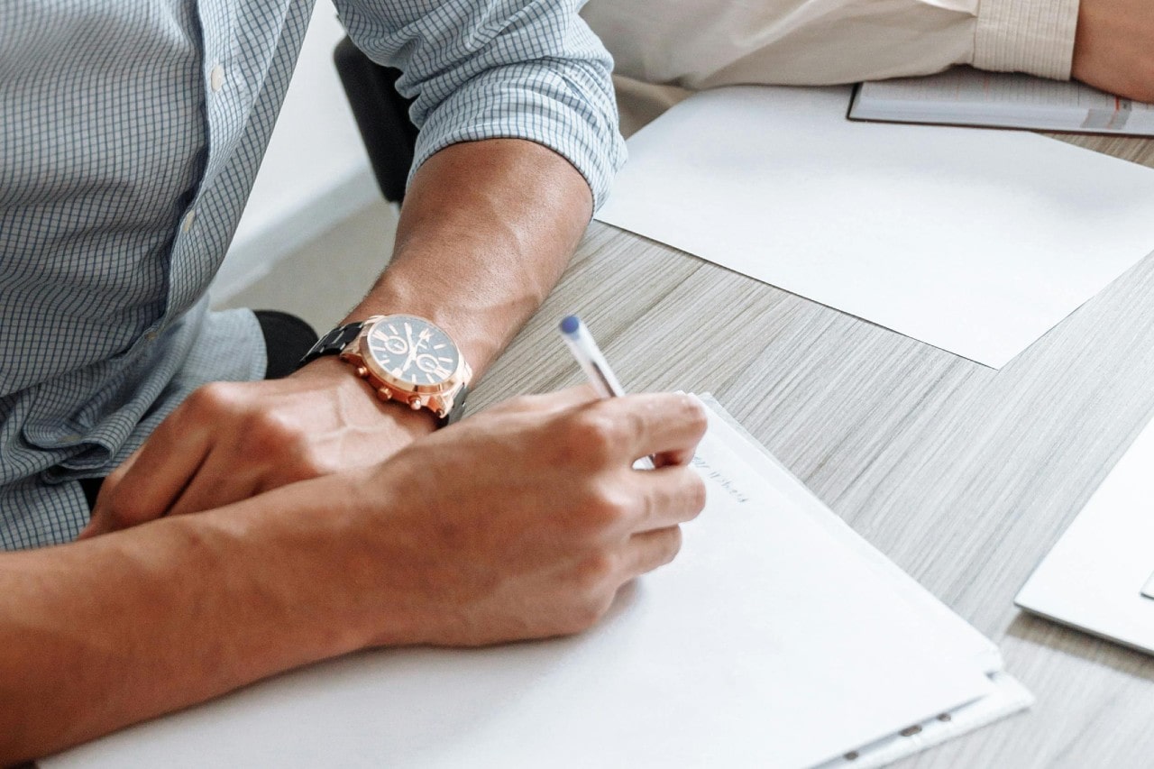 a man writing on a piece of paper and resting his arm–adorned with a rose gold watch