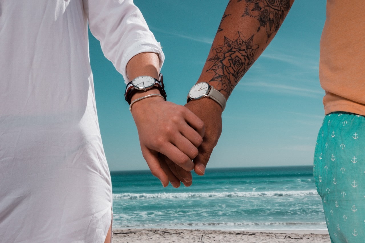 A close-up of a couple wearing luxury watches holding hands at the beach.