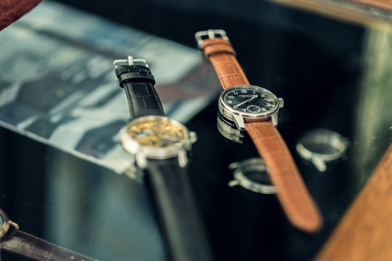 A close-up of two luxurious watches with leather bands displayed side-by-side on a dark tabletop.